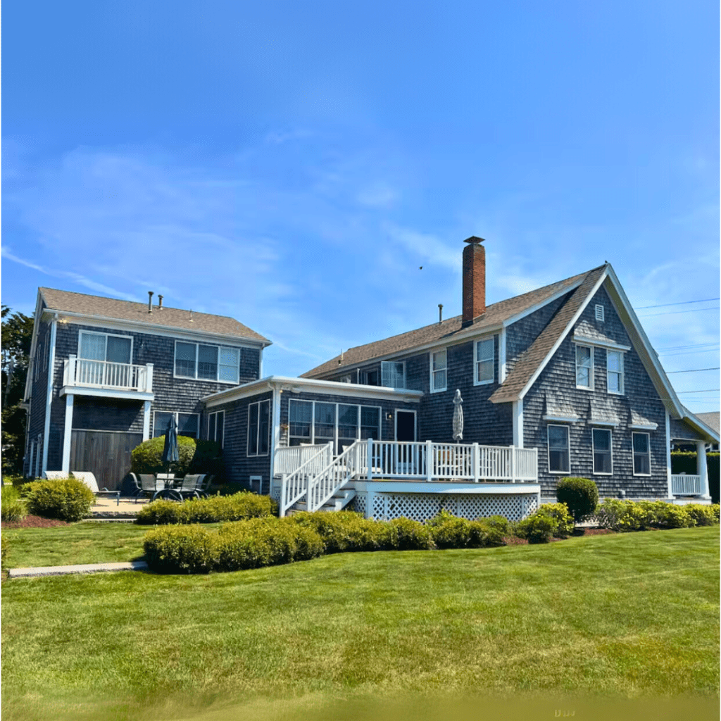 A large coastal home with a gray shingle exterior, featuring a wrap-around porch and multiple windows, set against a clear blue sky and green lawn.