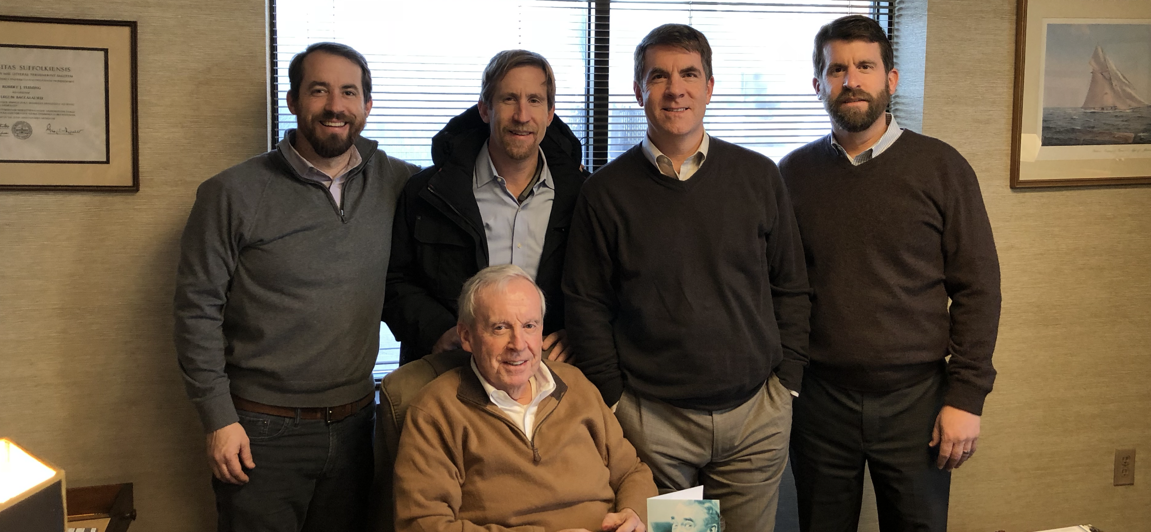 A group of five men, including one seated, poses together in an office setting with natural light coming through the windows.