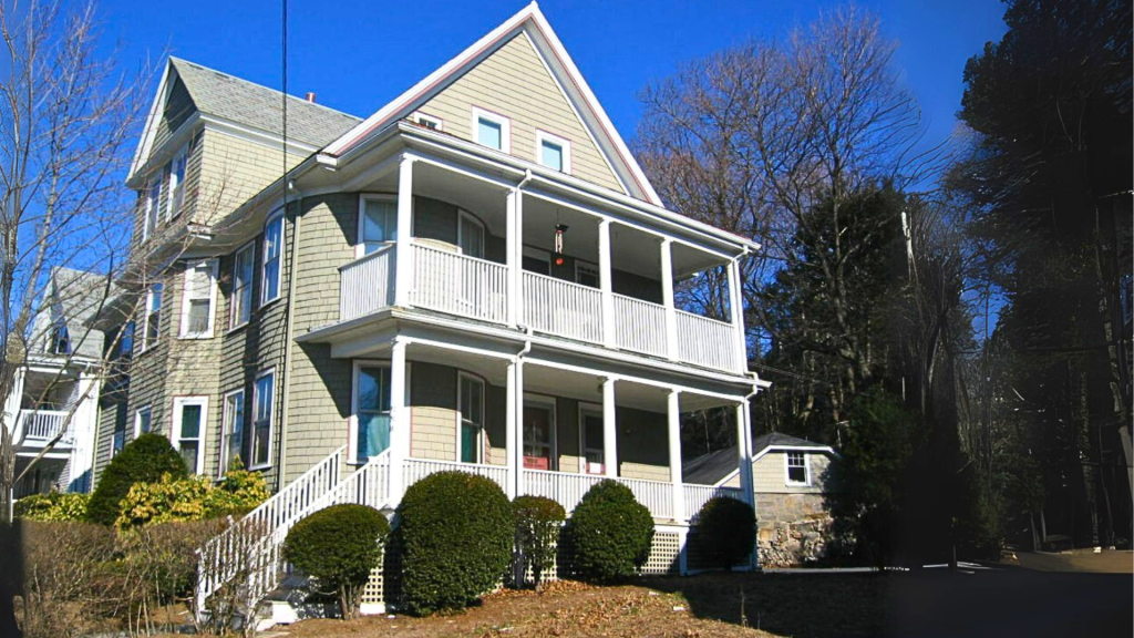 A multi-story residential building with a gabled roof and a balcony, surrounded by trees and shrubs under a clear blue sky.