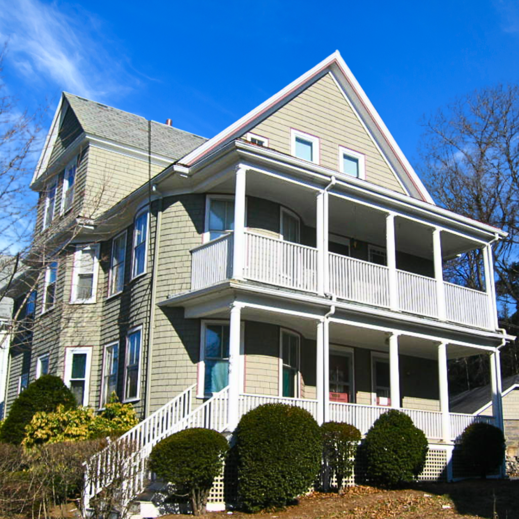 A large multi-family house with a prominent front porch and balconies, surrounded by well-maintained bushes.